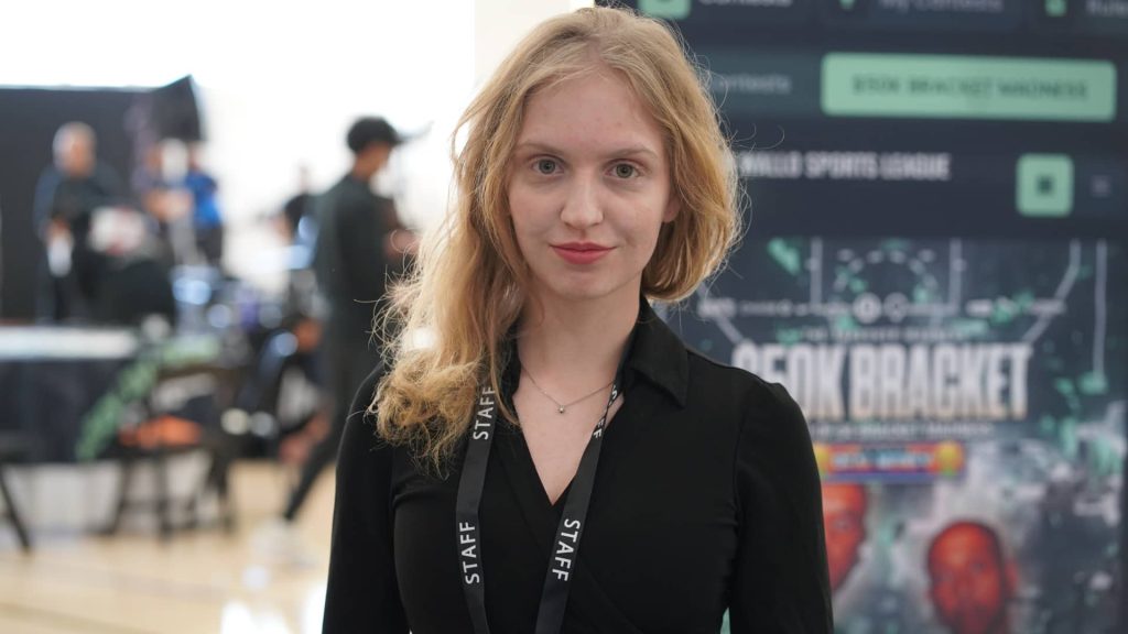 Female event staff member wearing a black outfit and “STAFF” lanyard, standing confidently at an indoor event with a branded display and attendees in the background.